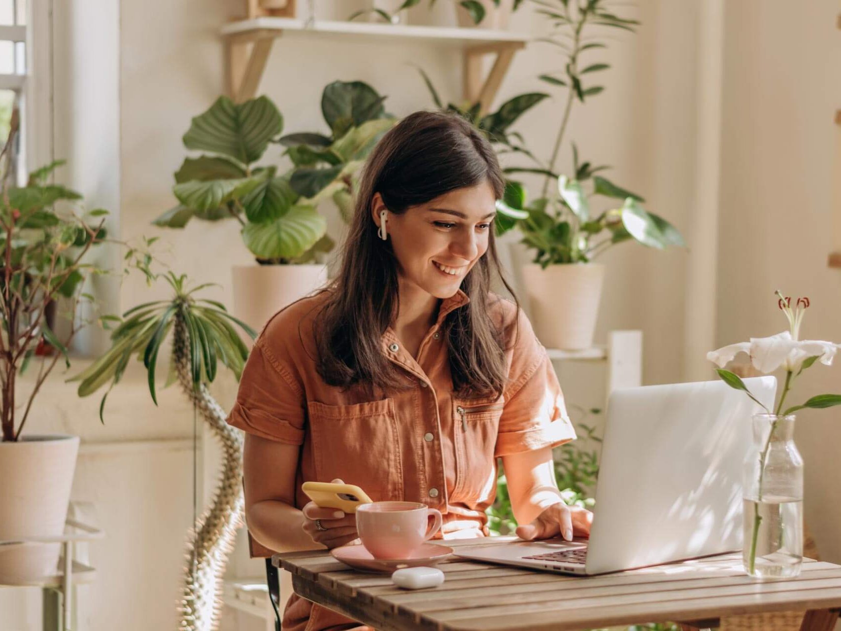 Eine Frau genießt eine Tasse Tee, während sie in einem gemütlichen, mit Pflanzen gefüllten Heimbüro an ihrem Laptop arbeitet.