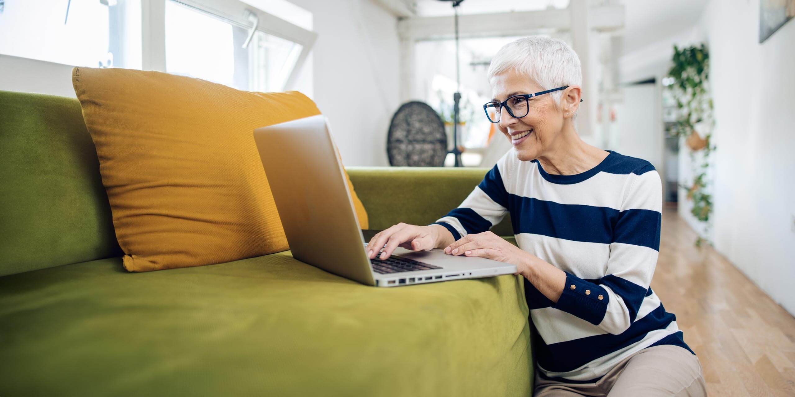 Ältere Frau mit Brille und stylischem Haarschnitt lächelt, während sie zu Hause auf einem grünen Sofa einen Laptop benutzt.