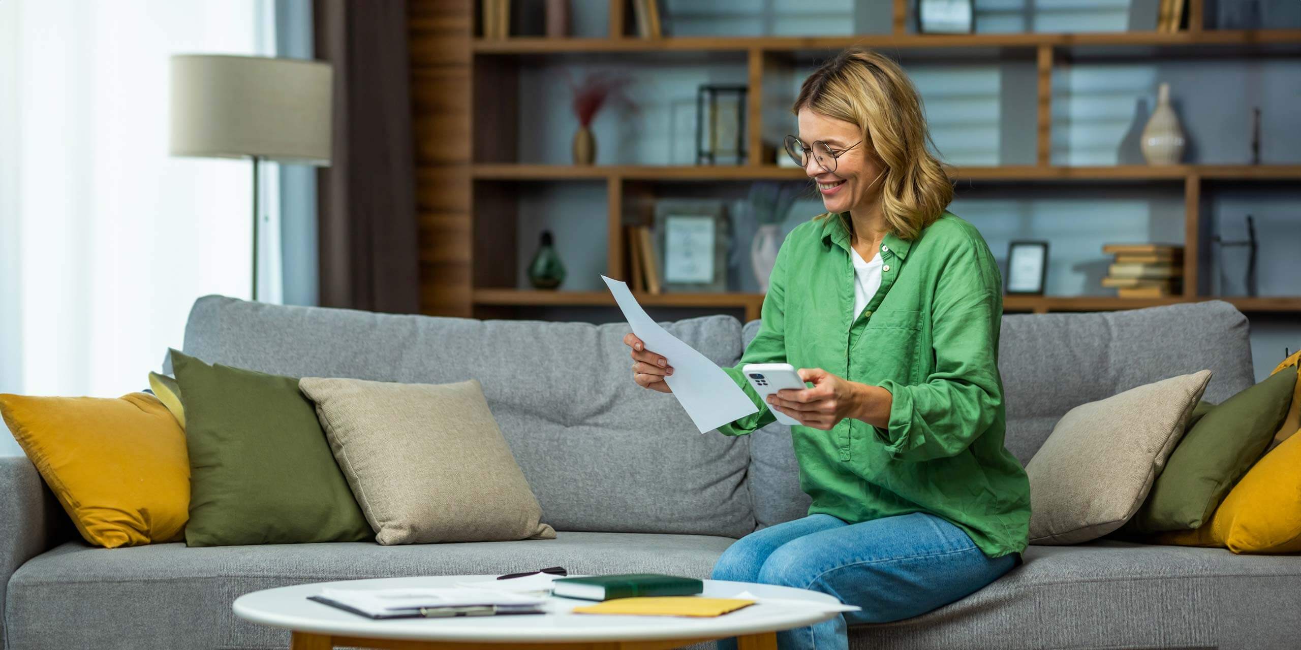Eine lächelnde Frau sitzt in einem gemütlichen Wohnzimmer auf einer Couch und betrachtet ein Dokument. In der anderen Hand hält sie ein Tablet-PC.