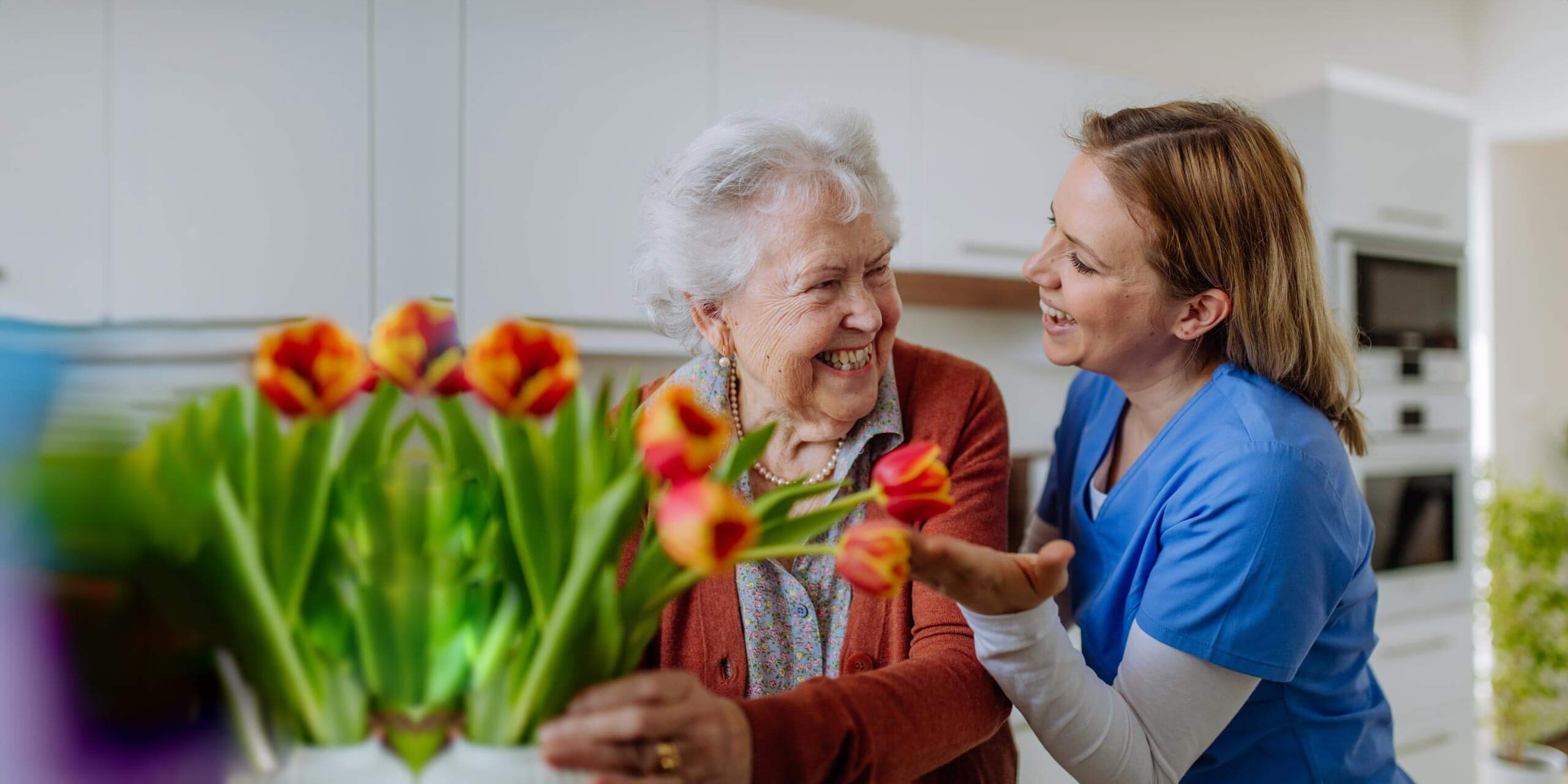 Eine fröhliche ältere Dame genießt ein nettes Gespräch mit einer jüngeren Dame, möglicherweise einer Pflegerin, in einer hellen Küche mit leuchtenden Tulpen, die der Szene einen Hauch von Wärme verleihen.